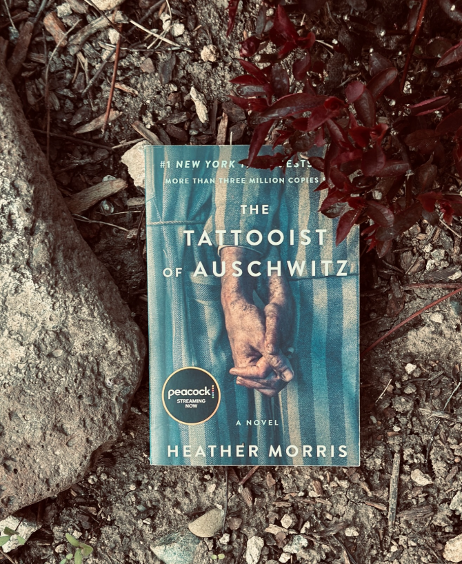A copy of "The Tattooist of Auschwitz" by Heather Morris lies on the ground among rocks, dirt, and reddish plants. The cover shows tattooed arms clasped together and includes a sticker about a Peacock streaming adaptation.