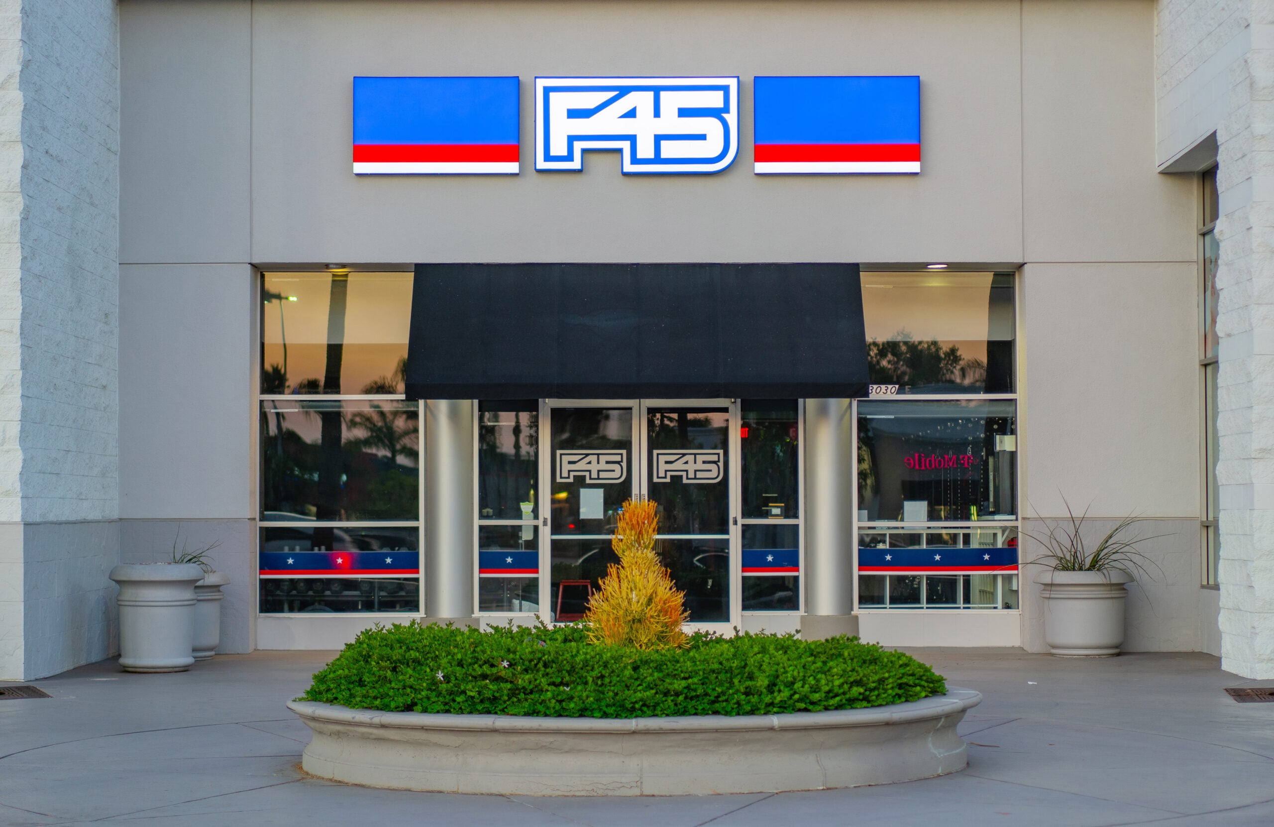 The image shows the exterior of an F45 gym with large windows, a blue and red F45 sign above the entrance, and a circular planter with greenery in front of the doors.