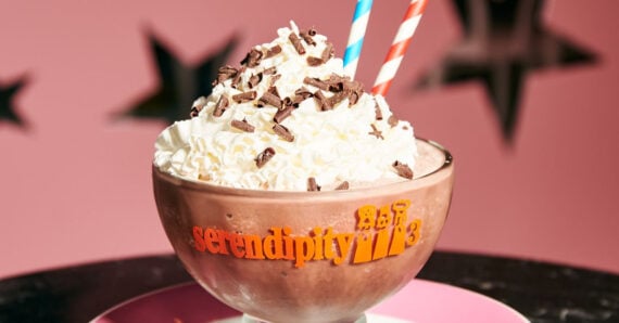 A large glass bowl labeled "serendipity 3" filled with a chocolate milkshake topped with whipped cream and chocolate shavings, with two striped straws, against a pink background with black star shapes.