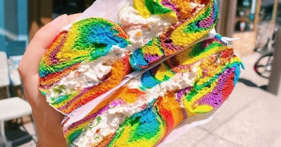 A hand holds a halved, vibrant rainbow bagel filled with cream cheese, with colorful swirls visible in the bread against an outdoor urban background.