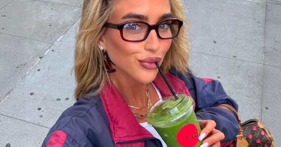 A woman with long blonde hair and glasses takes a selfie while drinking a green smoothie with a black straw. She is wearing a dark jacket with a maroon collar and holding a brown patterned bag. Pavement is visible in the background.