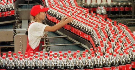 A factory worker in a red cap and overalls inspects rows of Coca-Cola bottles moving along a conveyor belt in a bottling plant.