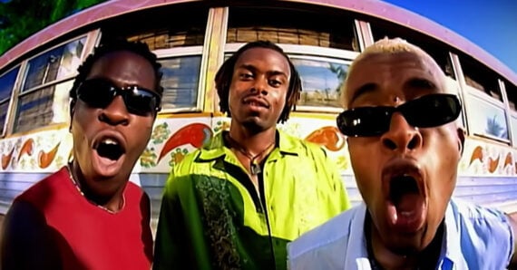Three men stand close to the camera in front of a colorful bus. They wear sunglasses and expressive faces, with one in a green shirt and the other two in red and light blue shirts, under a bright blue sky.