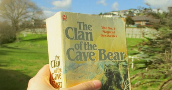 A hand holds a worn copy of "The Clan of the Cave Bear" book outdoors, with a green grassy yard, trees, fence, and houses in the blurred background under a partly cloudy sky.