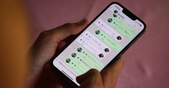 A person holds a smartphone displaying a WhatsApp chat screen with multiple green voice message notes in a conversation. The background is blurred in pink tones.