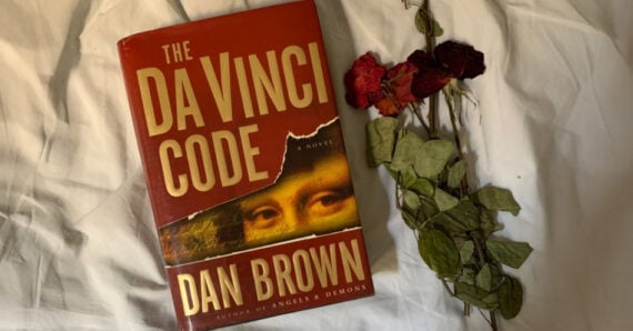 A copy of the book "The Da Vinci Code" by Dan Brown lies on a white fabric surface next to a small bundle of dried red roses and green leaves.