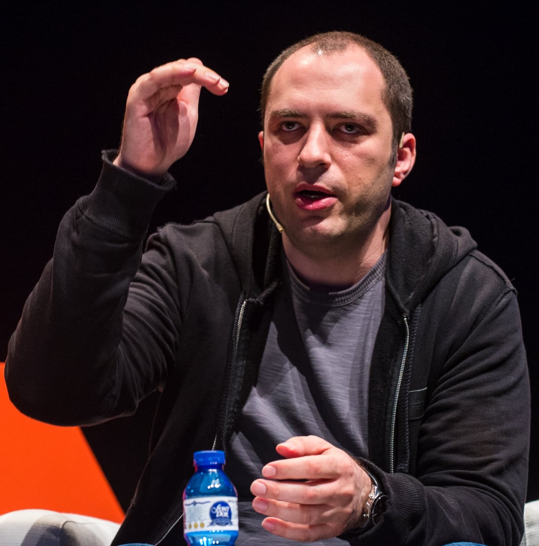 A man in a dark hoodie and gray t-shirt gestures with his hand while speaking. He is seated, with a blue water bottle in front of him, against a dark background with orange shapes.