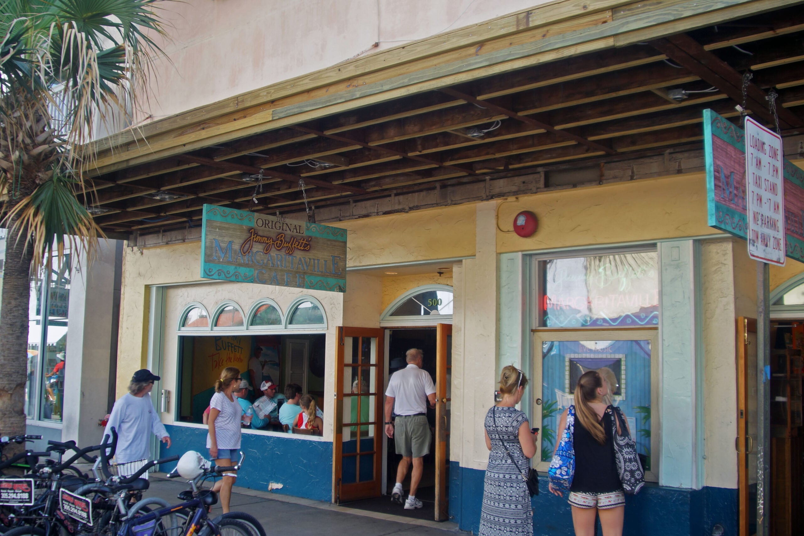 People stand outside and near the entrance of a casual beachside restaurant called "Original Longboards Margarita Bar & Grill" with bikes parked to the left and a palm tree nearby.