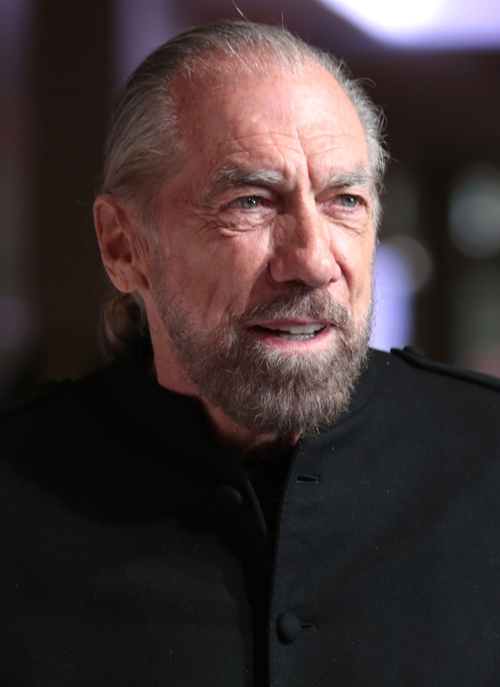 A man with gray hair and a beard, wearing a black jacket, is looking slightly to the side and smiling. The background is softly blurred with lights and indistinct shapes.