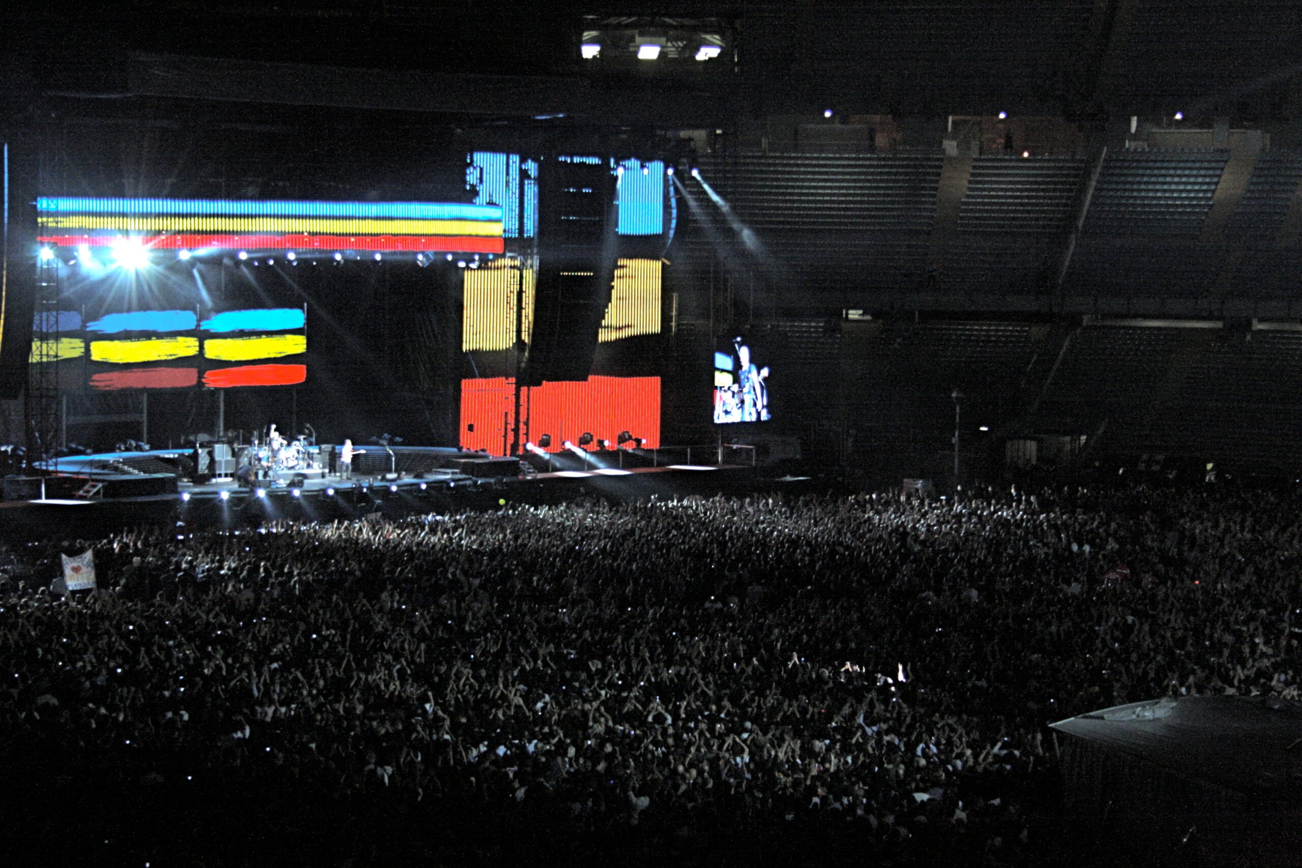 A large crowd at a concert in a stadium with colorful abstract visuals on a screen behind the stage and bright stage lights shining.