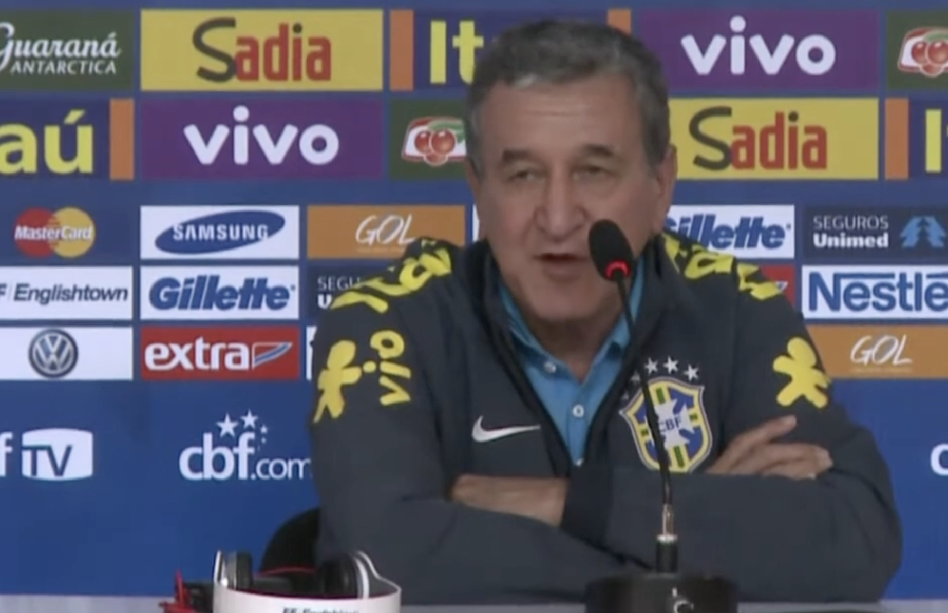 A man wearing a Brazil national football team jacket speaks at a press conference, sitting in front of a microphone with sponsor logos visible on the backdrop behind him.