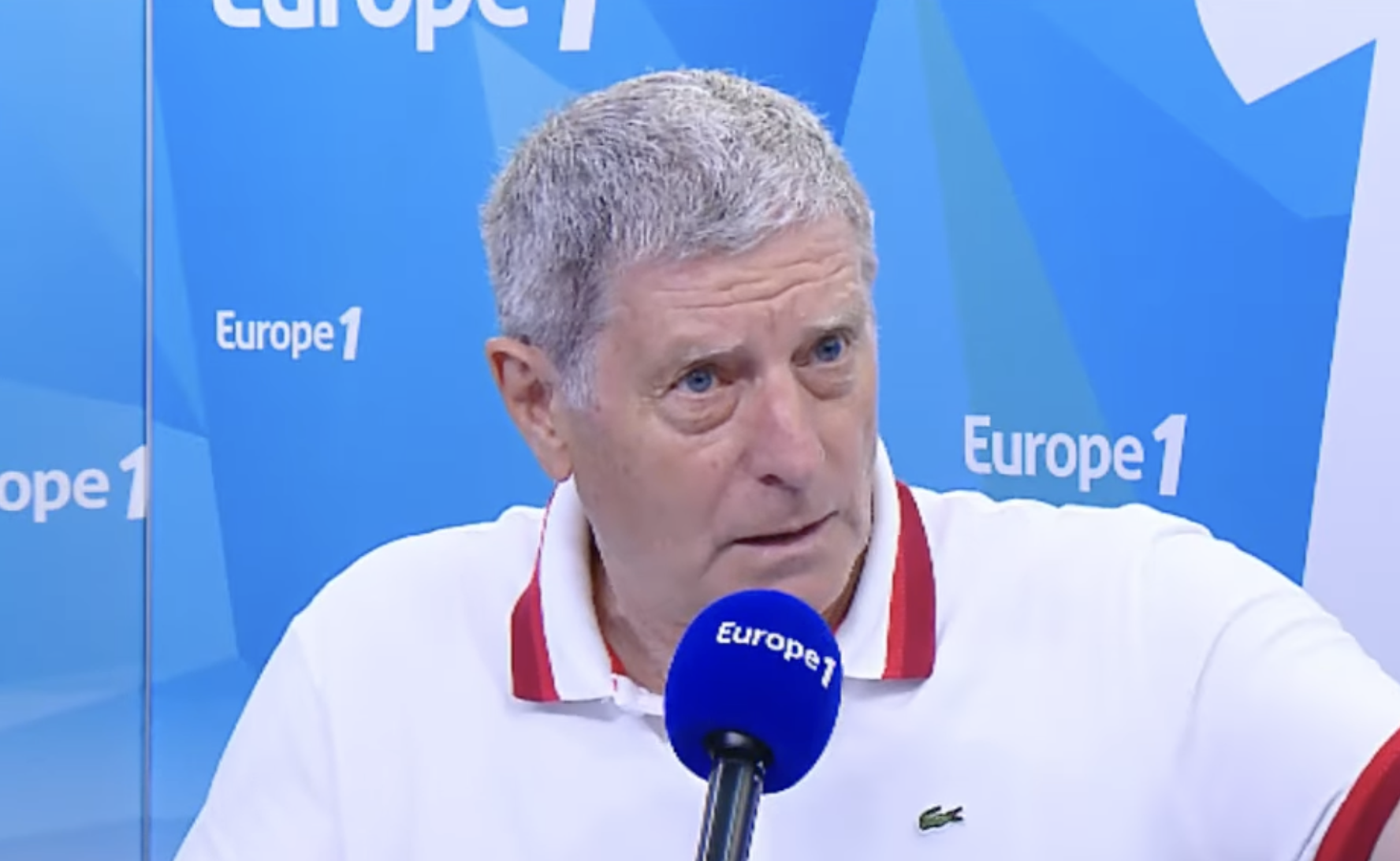 An older man with short gray hair, wearing a white polo shirt with red trim, speaks into a blue “Europe 1” microphone during a radio interview. The background displays the blue Europe 1 logo.