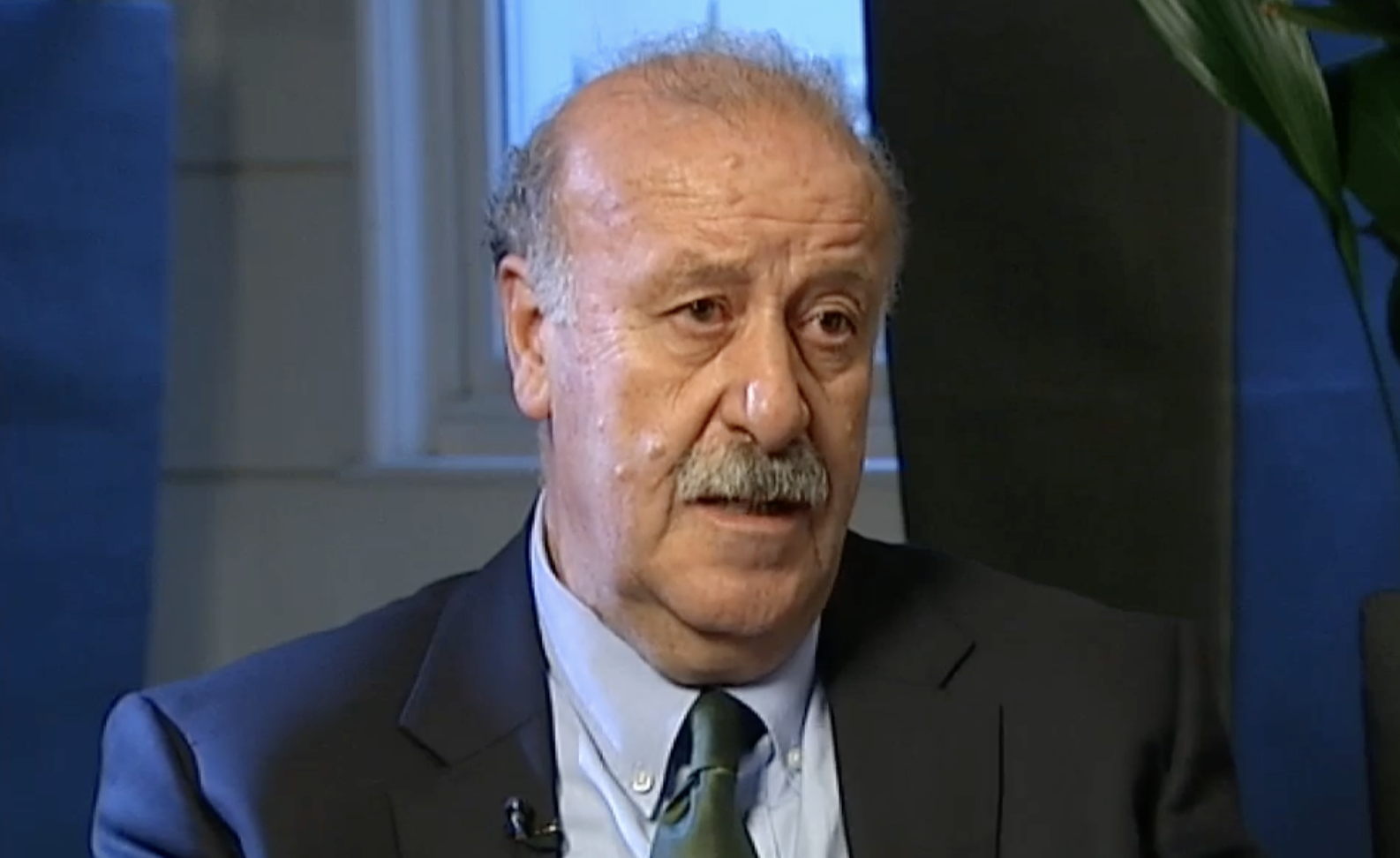 An older man with a mustache, wearing a suit and tie, is seated indoors. He appears to be speaking or in an interview. There is a window and a plant visible in the background.