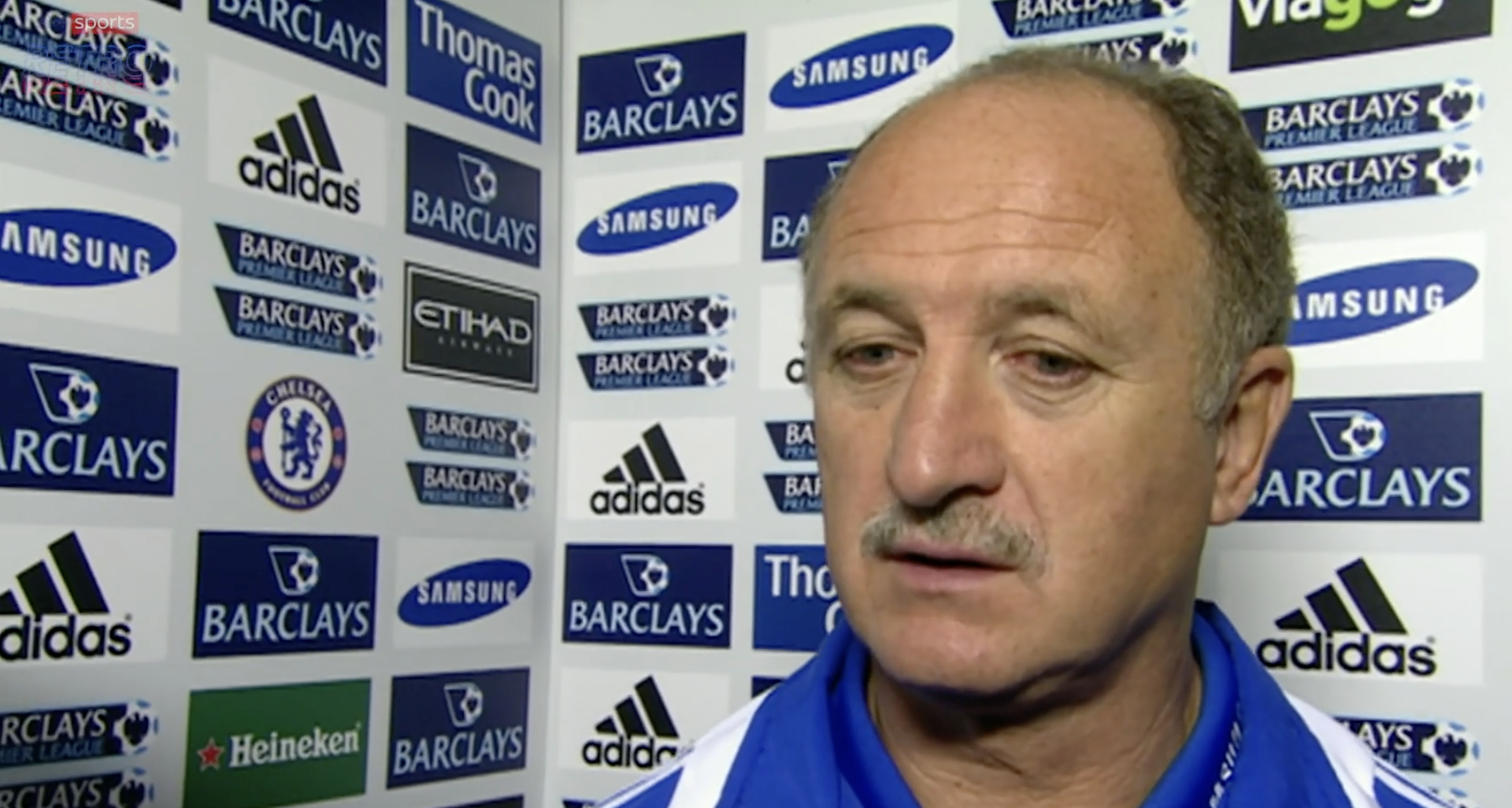 A man with a mustache wearing a blue and white sports jacket gives an interview in front of a backdrop covered with various sponsor logos, including Barclays, Adidas, Thomas Cook, and Chelsea FC.