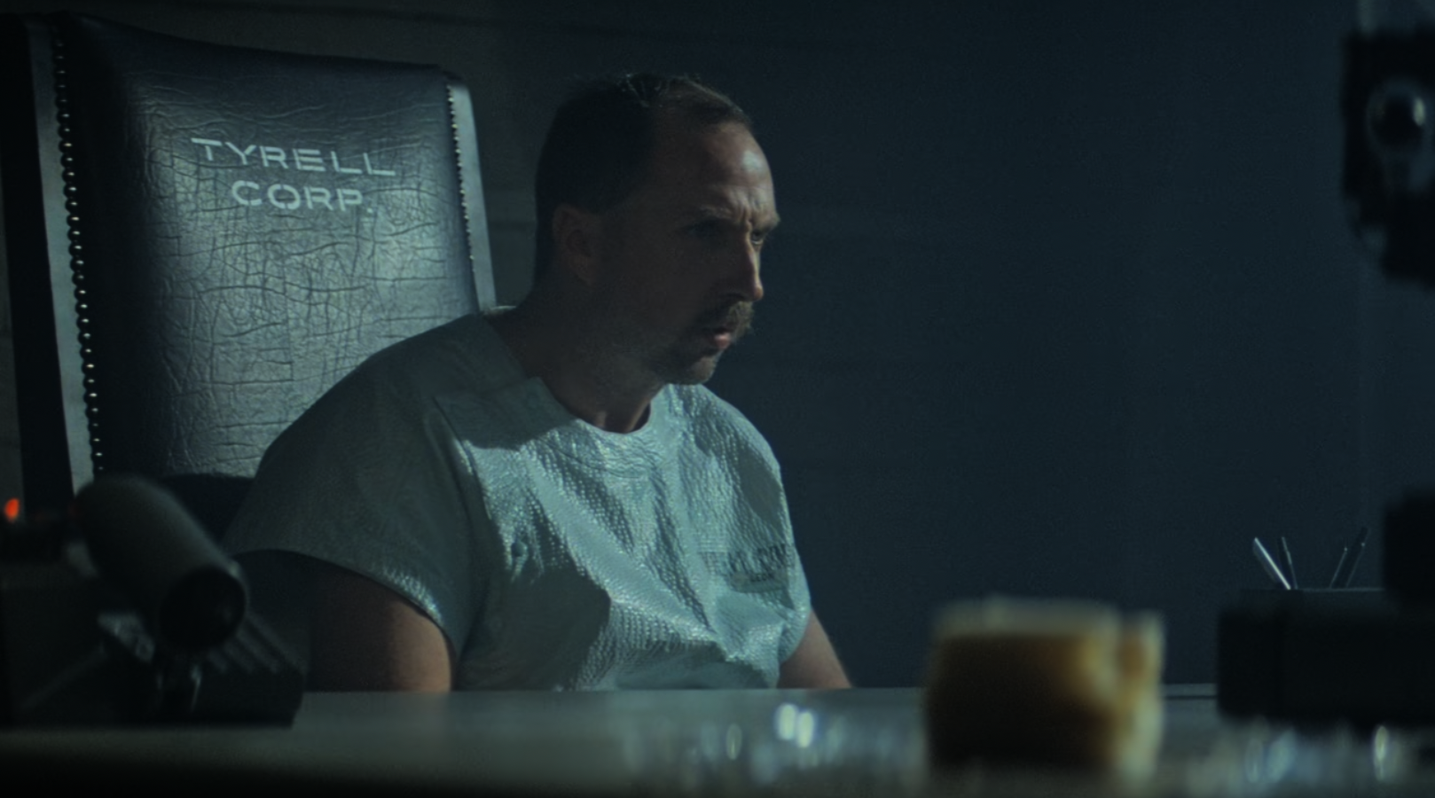 A man in a hospital gown sits in a dark room on a chair labeled "Tyrell Corp," looking tense and serious, with dim lighting and blurred objects in the foreground.