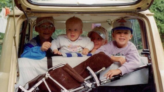 Four children smile and pose inside the open trunk of a car, with a folded stroller in front of them. The scene is outdoors, and the children appear happy and playful.