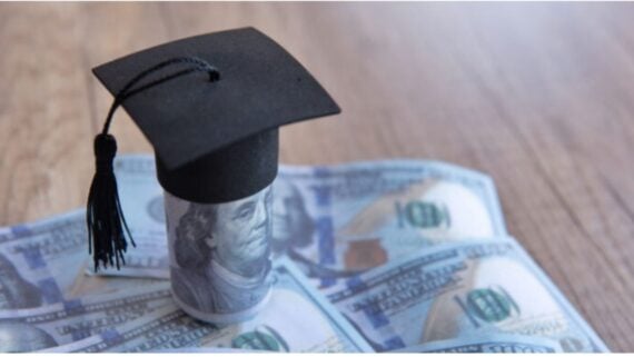 A graduation cap sits on a rolled-up U.S. hundred-dollar bill, surrounded by more hundred-dollar bills on a wooden surface, symbolizing the cost of education.