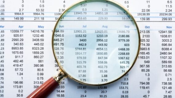 A magnifying glass focuses on numerical data in columns and rows on a printed financial spreadsheet or report, highlighting figures under the months of June, July, August, and September.