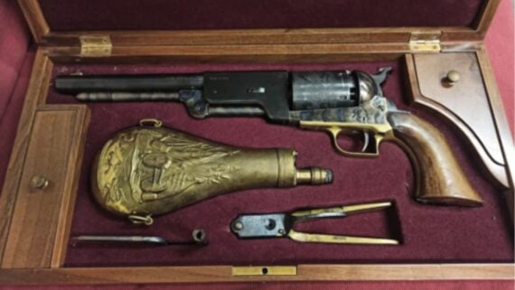 A vintage revolver in a wooden display case with a red velvet lining, accompanied by a brass powder flask, bullet mold, and cleaning tools, all arranged neatly in individual compartments.