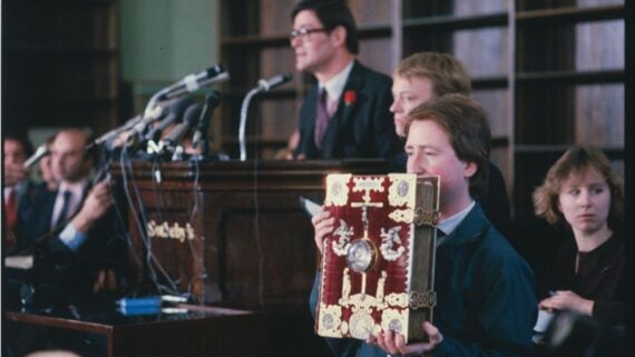 A person holds up an ornate, gold-embellished book in front of a seated audience at an auction, while several people speak at a podium surrounded by microphones. Shelves filled with books are visible in the background.