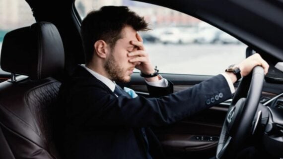 A man in a suit sits in the driver's seat of a car, resting his head in one hand with a stressed or frustrated expression. His other hand is on the steering wheel.