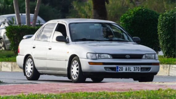 A silver, four-door Toyota sedan is driving on a street near greenery and bushes. The car has a blue-and-white license plate and its windows are slightly tinted.
