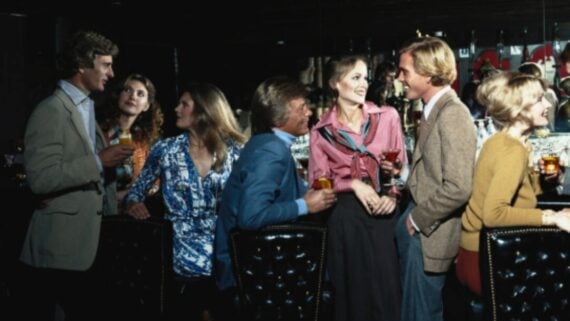 A group of well-dressed men and women socializing at a dimly lit bar, smiling and talking while holding drinks, with a row of bottles and glasses visible in the background.