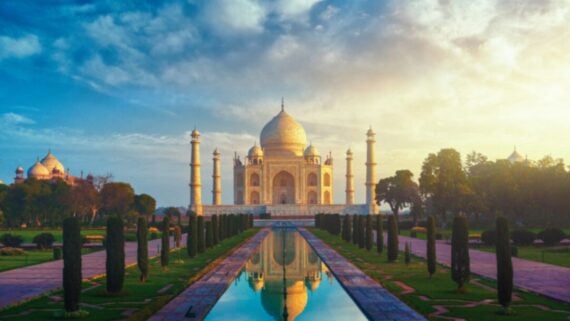 The Taj Mahal in India at sunrise, with its white marble dome and minarets reflected in a long, narrow pool, surrounded by lush gardens and a dramatic sky with clouds.