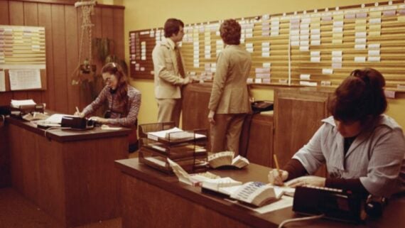 Two women work at desks with rotary phones and paperwork, while two men in suits stand at a large wall of schedules or files in a retro, wood-paneled office with yellow walls.