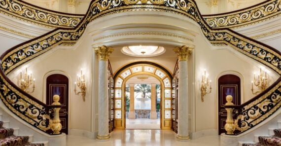 Grand entrance hall with an ornate double staircase, gold and black railings, marble columns, elegant wall sconces, and a large arched doorway with intricate glasswork leading to a sunlit area outside.