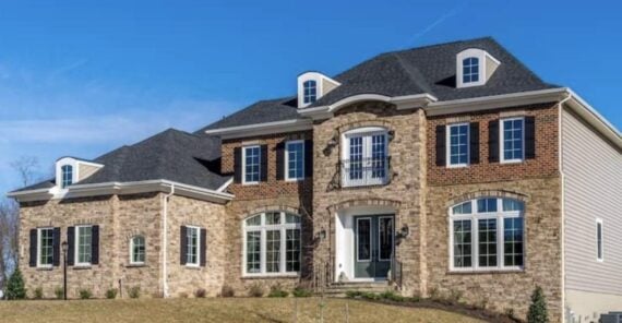 A large two-story house with brick and siding exterior, black shutters, and multiple windows. The home features a grand entrance with a small balcony above the front door and a sloped yard in the foreground.