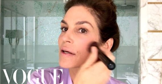 A woman with dark hair tied back applies makeup to her cheek using a brush in a bright bathroom. The Vogue logo appears in the lower left corner of the image.