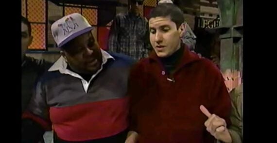 Two men stand close together indoors; one wears a multicolored rugby shirt and a baseball cap, while the other wears a red pullover. They appear to be talking, with the man in red gesturing with his hand.