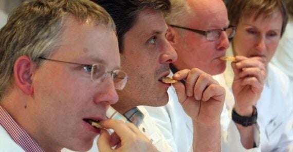 Four people in white lab coats are sitting in a row, closely tasting crackers or biscuits, suggesting a food tasting or quality control session.