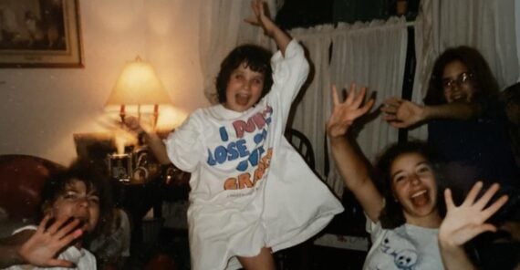 Four children are indoors, smiling and waving their hands toward the camera. One child stands in the center, wearing a large white t-shirt and appearing excited. A lamp and curtains are visible in the background.