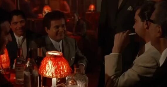Four men in suits sit around a dimly lit table with red lamps, drinks, and bottles, appearing to enjoy a lively conversation in a bar or nightclub setting.