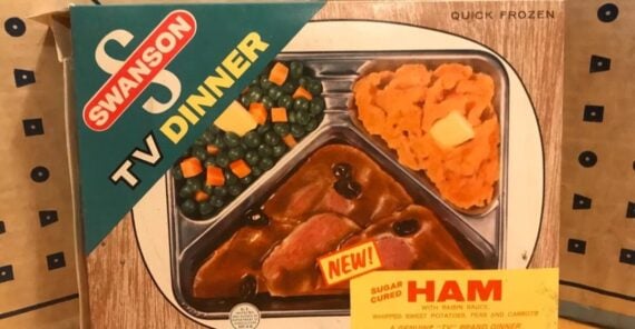 Vintage Swanson TV Dinner box featuring a photo of a meal with ham in sauce, sweet potatoes, peas, and carrots. The packaging is labeled "Sugar Cured Ham" and "NEW!" with a 1950s-style design.