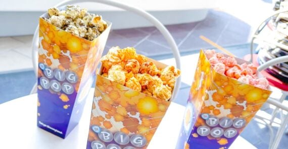 Three tall, colorful popcorn boxes labeled "Big Pop" sit on a white table, each filled with different flavored popcorn: one brown, one orange, and one pink.