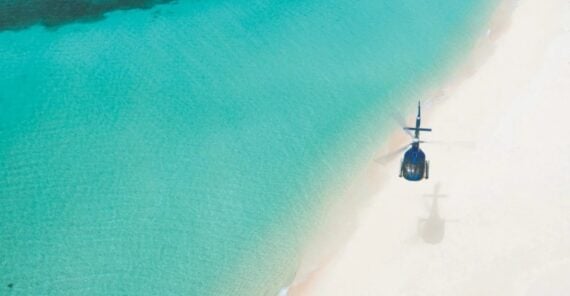 A blue helicopter flies over a pristine beach, casting a shadow on the sand where turquoise water meets the shore.
