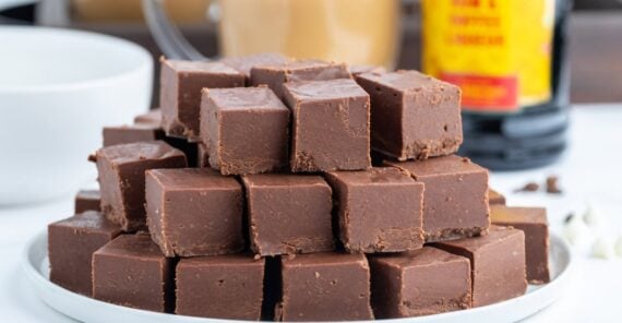 A white plate stacked high with neatly cut squares of rich, creamy chocolate fudge, set on a white surface with a blurred mug and bottle in the background.