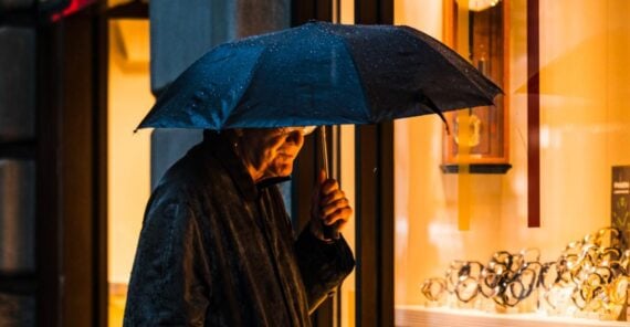 An older man stands outside in the rain under a black umbrella, looking into a brightly lit shop window displaying watches and jewelry.