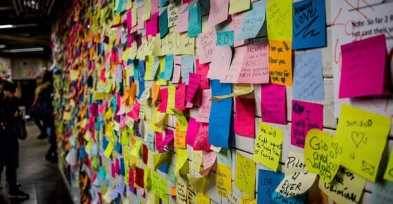A wall covered in colorful sticky notes filled with handwritten messages, drawings, and doodles, with some people standing and observing the notes in the background.