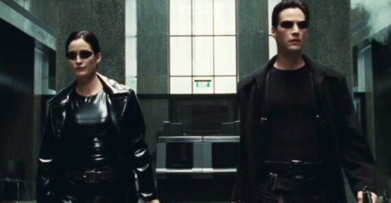 Two people wearing black leather outfits and sunglasses walk confidently through a modern, industrial hallway with green-tinted walls and glass panels.