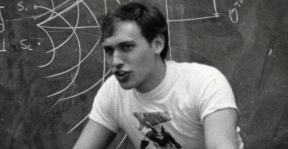 A young man wearing a graphic t-shirt speaks animatedly in front of a chalkboard covered with diagrams and writing.