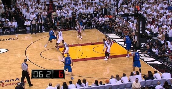 NBA basketball game in action; one player dribbles near the three-point line as defenders close in. The court is packed with players and spectators. A shot clock displays 6 seconds remaining.