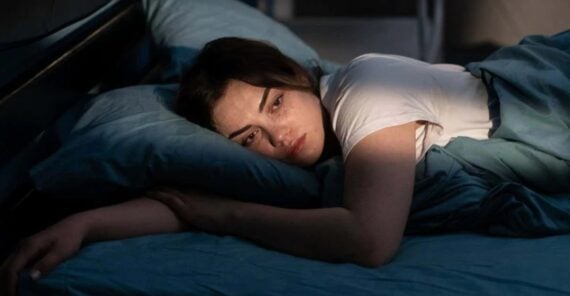 A woman lies in bed on her side, resting her head on a blue pillow and looking ahead with a tired or pensive expression. Sunlight partially illuminates her face and arm.