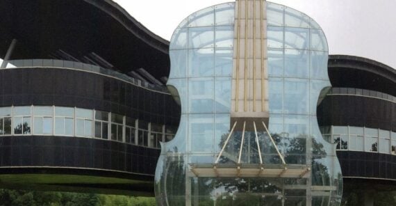 A modern building with a large glass entrance shaped like a violin, connected to a curved, black-panel structure with rows of windows. Trees and sky are reflected on the glass.