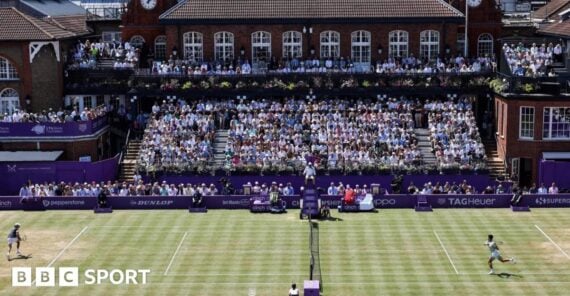 Two tennis players compete on a grass court in front of a large seated crowd at an outdoor stadium, with historic red-brick buildings in the background. The BBC Sport logo is visible in the corner.