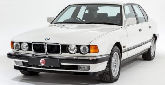 A white classic BMW sedan, likely from the late 1980s or early 1990s, is parked in a studio setting with a clean white background. The car features round headlights and signature kidney grilles.