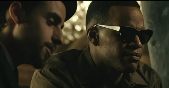 Two men sit closely together in a dimly lit room; one wears sunglasses and a neutral expression while the other leans in, partially smiling, both appearing deep in conversation.